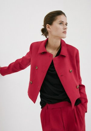 Mujer con chaqueta y pantalones rojos a medida y top negro, posando con un brazo extendido contra un fondo blanco.