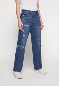 Persona vestida con jeans rectos desgastados azules con rotos en las rodillas y zapatillas blancas, de pie contra un fondo sencillo.