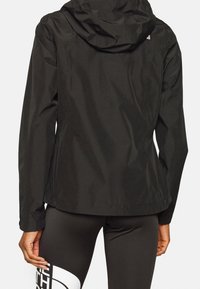 Persoon in een zwarte hoodie en zwarte legging met een groot wit logo op de dij, gezien vanaf de achterkant.