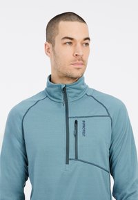 Pullover in pile color turchese con mezza zip, alto colletto e tasca a zip sul petto. Presenta un tessuto testurizzato e cuciture a contrasto.