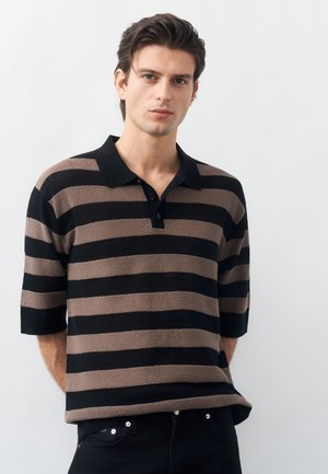 Νέος άνδρας με σκούρα μαλλιά, ντυμένος με κοντομάνικη πόλο μπλούζα με οριζόντιες ρίγες σε μαύρο και καφέ χρώμα, στέκεται μπροστά σε απλό φωτεινό φόντο.
