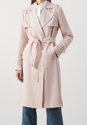 Frau trägt einen hellrosa Trenchcoat mit Gürtel und Knopfdetails, dazu blaue Jeans und ein weißes Hemd mit Kragen.