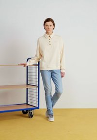 Beige hosszú ujjú pólóing gombos nyakával, világoskék farmernadrággal és szürke tornacipővel párosítva. Egy kék fém kocsi mellett áll, sárga padlón.