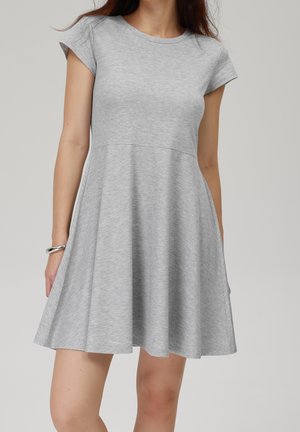 Femme portant une robe trapèze gris clair à manches courtes, longueur genou, avec un col rond et un bracelet argenté au poignet gauche.