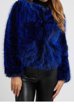 Femme portant une veste en fausse fourrure rayée bleue et noire à manches longues et un pantalon en cuir noir, debout devant un fond uni.
