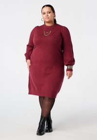 Maroon gebreide jurk tot op de knie met een visgraatpatroon, ballonmouwen en geribbelde manchetten. Draagt zwarte enkellaarzen en gelaagde kettingen.