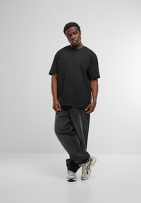 Zwarte oversized t-shirt met korte mouwen, gecombineerd met losse donkergrijze jeans en metallic sneakers, tegen een lichtgrijze achtergrond.
