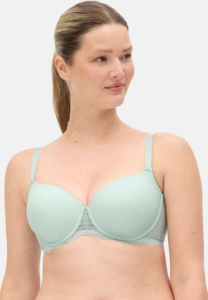 Mujer con cabello castaño claro recogido en una cola de caballo, lleva un sujetador acolchado verde claro con detalles de encaje, mirando ligeramente hacia la derecha.