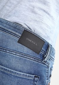 Jeans de mezclilla en azul claro con una superficie texturizada. Presenta un parche de cuero negro etiquetado como "DIESEL" en la parte posterior de la cintura. Detalles de costura visibles.