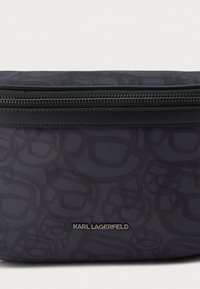 Svart stoffpose med overlappende mønster av mørke solbriller, sølvglidelås og sentrert "KARL LAGERFELD" metalllogo.