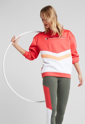 Felpa rossa e bianca con chevron arancioni e logo "TOMMY SPORT", abbinata a leggings verdi con accenti rossi e bianchi.