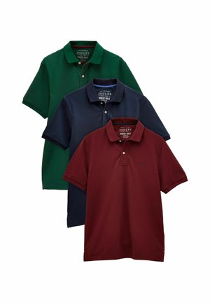 Man staat met handen in de zakken, draagt een bordeauxrode polo met korte mouwen en lichtbeige broek tegen een effen beige achtergrond.
