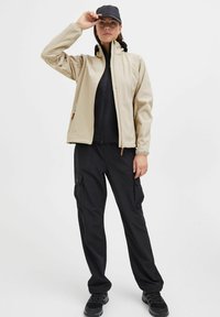 Beige softshell jacket with high collar and zippered pockets, layered over a black top, paired with black cargo pants and black sneakers.