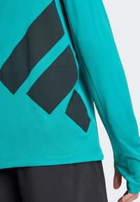 Długi rękaw sportowa bluzka z turkusowej siateczki z czarnymi geometrycznymi akcentami. Posiada dopasowany krój i otwory na kciuki przy mankietach.