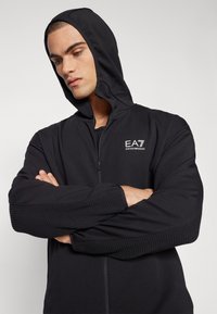 Musta vetoketjulla varustettu huppari, joka on valmistettu sileästä kankaasta. Sen hihoissa on tekstuuripainatus ja rinnassa EA7-logo. Huppu tekee siitä urheilullisesti istuvan.