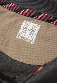 Label met "J&JOY" logo op beige stof, met een zwart en roze patroonrand. Gladde textuur en afgeronde hoeken.