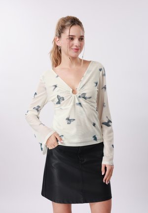 Jonge vrouw met een licht, met vlinders bedrukt shirt met lange mouwen en een zwarte leren rok, staand met één hand in de zak.