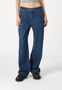 Vaqueros de denim azul holgados con cintura alta, que cuentan con bolsillos frontales, costuras naranjas y un ajuste relajado. Combinados con zapatos negros.