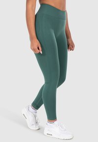 Leggings verdi a vita alta realizzati in tessuto elasticizzato, con una texture liscia, cuciture laterali e abbinati a scarpe da ginnastica bianche.