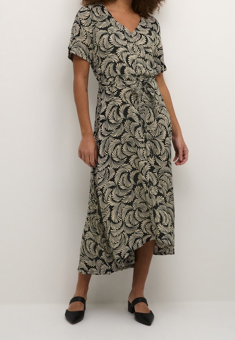 Schwarzes Kleid mit weißem Blattmuster, kurzen Ärmeln, V-Ausschnitt, Taillengurt und asymmetrischem Saum. Getragen mit schwarzen Blockabsatzschuhen.
