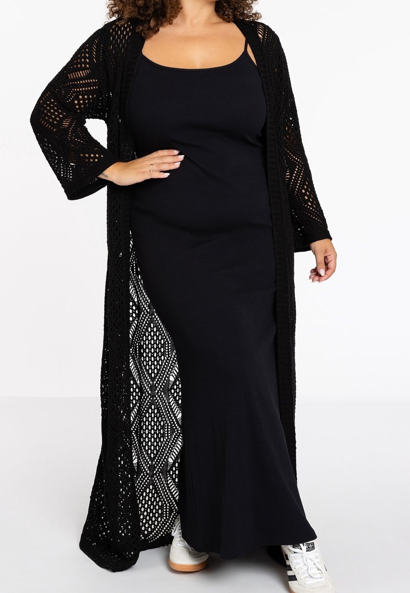 Femme portant une longue robe noire sans manches et un cardigan en maille noire ouverte, associée à des baskets blanches, debout avec une main sur la hanche.