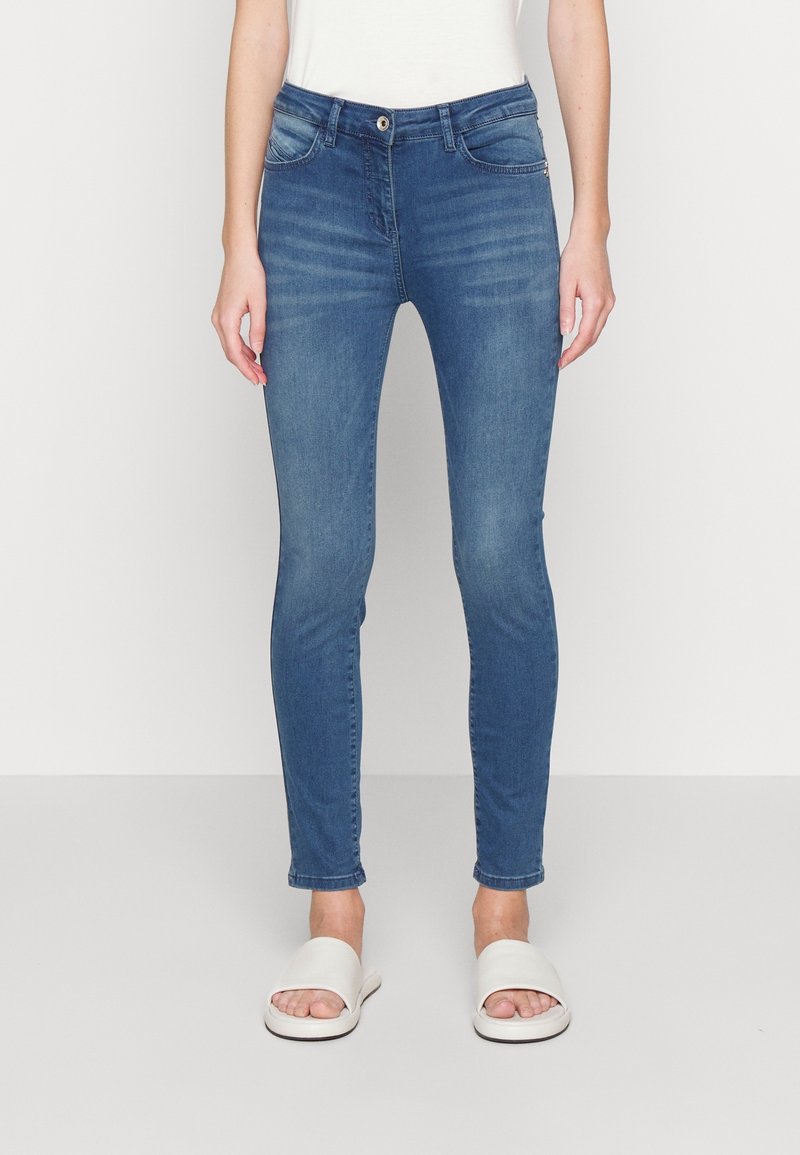 Jeans de denim ajustados en azul medio con ligero desgaste, dos bolsillos delanteros y cierre de botón en la parte frontal. Combinados con sandalias deslizables blancas.