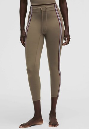 Slim-fit beige legging met zijstrepen en een trekkoord in de taille, gedragen door een staand persoon zonder schoenen tegen een effen lichte achtergrond.