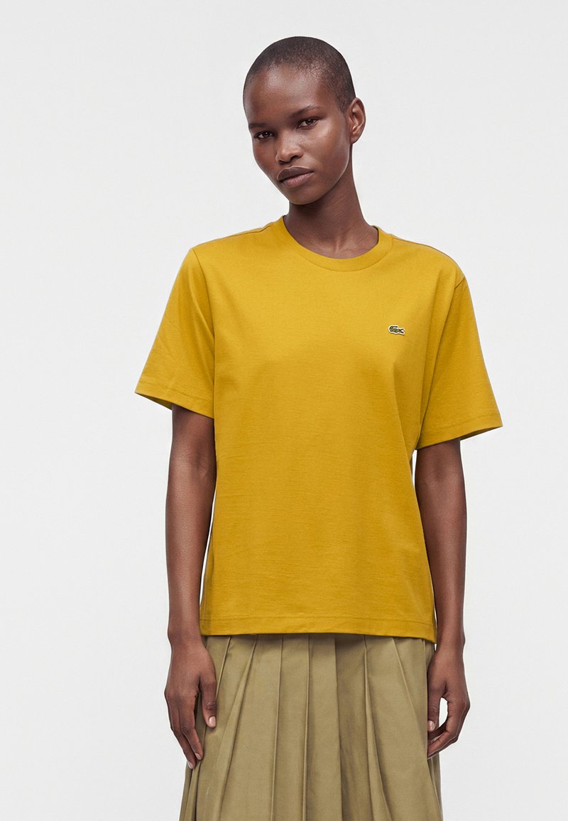 Personne aux cheveux courts portant une chemise jaune moutarde à manches courtes et une jupe plissée beige, debout devant un fond blanc uni.