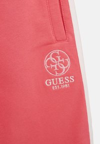 Pantaloni della tuta in cotone rosa con tasche laterali, dettagli bianchi e logo ricamato "GUESS EST.1981". Texture liscia e vestibilità comoda.