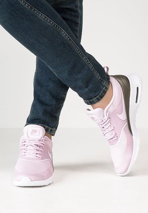 Personne portant des baskets Nike rose clair avec des accents foncés et un jean bleu foncé, un pied croisé sur l'autre sur un fond uni.
