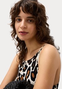 Mouwloos topje met een luipaardprint in zwart, bruin en roomkleurig. Het model heeft krullend haar en houdt een gestructureerde zwart gevlochten tas vast.