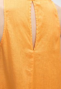 Οπίσθια όψη μιας αμάνικης πορτοκαλί λινής μπλούζας με V-σχήματος άνοιγμα στο επάνω κέντρο.