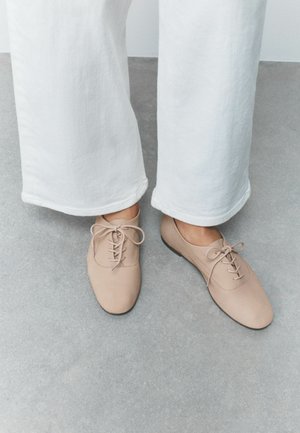 Persoon die beige veterschoenen en wijde witte broek draagt en op een grijze vloer staat.