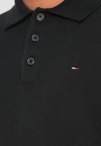 Mustast tekstuurse kudum materjalist polo särk. Omab kolme musta nuppu ning väikest sinist, valget ja punast värvi logo.