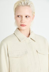 Beige teksturert jakke med kragehals, to brystlommer og lange ermer, mot en lys bakgrunn.