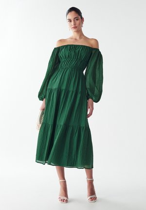 Vestido verde off-the-shoulder con mangas largas abullonadas, tela texturizada, diseño en capas, de longitud media, combinado con tacones blancos con tiras y un clutch.