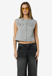 Sivi traperi kratki prsluk s ravnim krojem, s kopčama na dugme i dva džepa na prsima, uparen s širokim crnim trapericama.