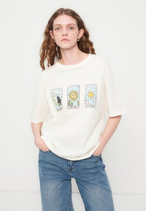 Mladi žena s valovitom kosom nosi bijelu majicu s motivima tarot karata: Mjesec, Sunce i Zvijezda, u kombinaciji s plavim trapericama.