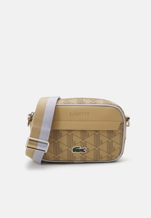 Sac bandoulière beige avec un motif géométrique, arborant un logo Lacoste, des détails en métal argenté et une bandoulière rayée réglable.