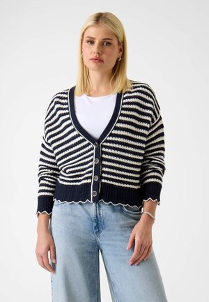 Blonde vrouw draagt een navy blauw-wit gestreepte cardigan met knopen over een wit shirt, gecombineerd met lichtblauwe high-waisted jeans, staand tegen een witte achtergrond.