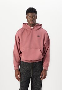 Haut à capuche surdimensionné rose avec logo noir, poche avant et clous décoratifs le long des manches. Associe à un pantalon noir texturé présentant des fissures.