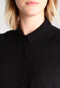 Chemise noire à boutons avec col classique, texture lisse, manches courtes et une poche poitrine avec un détail subtil de couture.