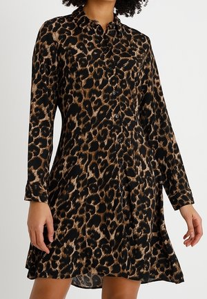 Kobieta ubrana w sukienkę na guziki z długim rękawem, w brązowo-czarny wzór w cętki lamparta.
