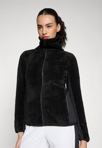 Jachetă din fleece neagră cu guler înalt, față texturată, panouri laterale negre contrastante, închidere cu fermoar frontal și buzunare laterale.