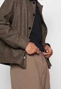 Veste en laine marron, texturée, avec un col, des boutons noirs et des poches latérales, portée sur un haut noir et un pantalon beige clair.