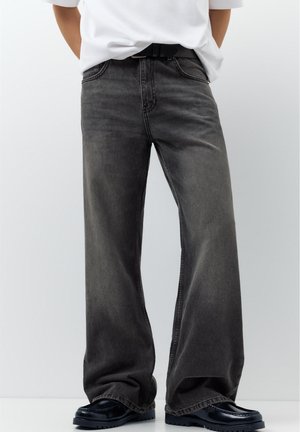 Person iført mørkegrå vidde jeans med sort bælte, hvid indstukket skjorte og sorte loafers, stående mod en ensfarvet baggrund.