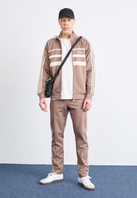 Bruin trainingspak met witte strepen, met rits jack en bijpassende broek. Gecombineerd met een witte t-shirt en witte sneakers. Zwarte pet en tas.