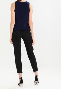 Marinefarbener ärmelloser Oberteil, kombiniert mit schwarzen, tapered Hosen. Schwarze Absätze mit weißen Punkten und einer Schleifen-Details am Knöchel. Einfaches und figurbetontes Design.