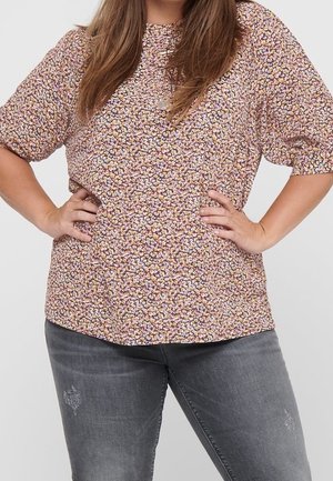 Mujer con cabello largo y castaño, que lleva una camiseta de flores de manga corta y vaqueros grises desgastados, con las manos en las caderas, sobre un fondo liso.