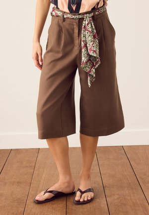 Kvinde iført brune, brede cropped bukser med et mønstret stofbælte, en flerfarvet top og mørkebrune flip-flops, stående på et trægulv.